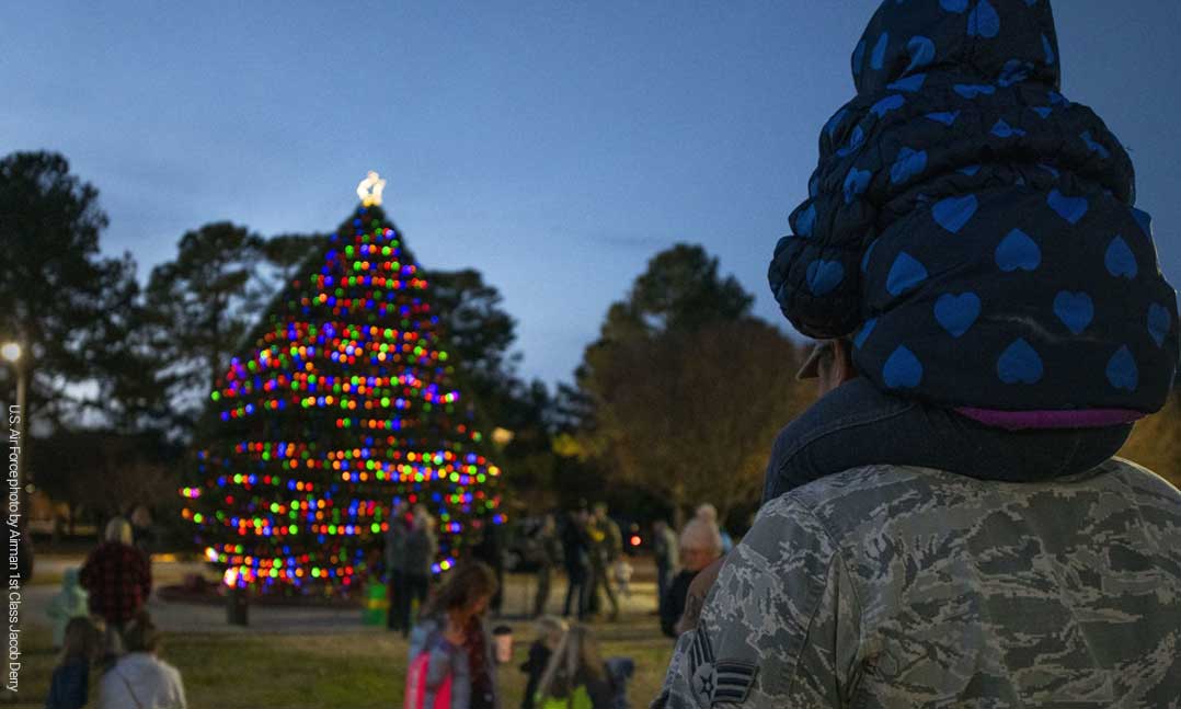 Soldier holds his child on his shoulders while standing with a crowd in front of a holiday tree with lights