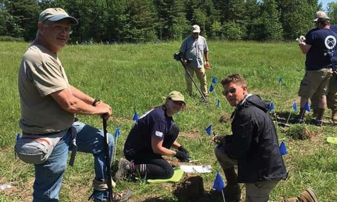 Army National Guard veteran Sandra Johnson, center, kneels on the ground to list archaeological finds on a roster using a GPS locator in June 2019 in Saratoga Springs, N.Y