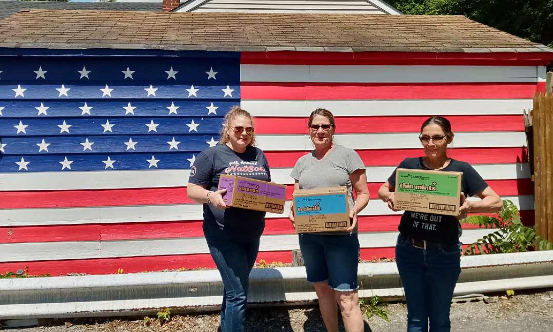 VFW Department of New York Service Officer Maddie Fletcher stands with two friends holding boxes in front of a US flag